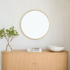 a round mirror on the wall above a wooden cabinet with a bowl and plant next to it