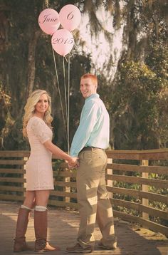 a man and woman holding onto pink balloons while standing on a wooden bridge with trees in the background