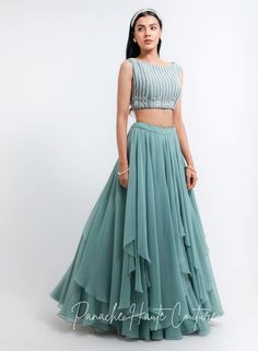 This exquisite ensemble features a stunning sage green tiered skirt crafted from lightweight georgette fabric, creating a flowy and airy silhouette. The skirt boasts multiple tiers that cascade gracefully down to the ankle, adding a touch of elegance to the overall look. Paired with the skirt is a beautiful blouse made from raw silk, adorned with intricate embroidery work using pearls, glass beads, and sequins petals. The embroidery adds a luxurious and glamorous touch to the blouse, making it p Georgette Skirt And Crop Top, Sage Green Skirt Outfit, Skirt And Crop Top Indian, Indowestern Dresses, Indowestern Gowns, Western Gowns, Indo Western Gown, Skirt And Crop Top, Lehenga Saree Design