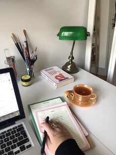 a person writing on a notepad next to a laptop computer and coffee cup with pencils in it