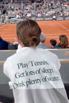 a woman sitting in front of a tennis court wearing a white shirt that says play tennis get lots of sleep, drink plenty of water