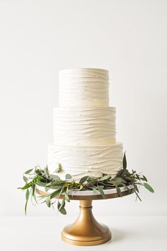 a white wedding cake with greenery on the top and bottom tier, sitting on a gold pedestal