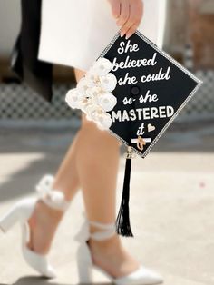 a woman in white high heels holding a black and white graduation cap with flowers on it