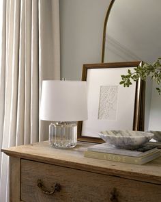 a wooden dresser topped with a lamp next to a mirror and a framed picture on top of it