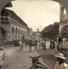 an old time photo of people and horses on the street with buildings in the background