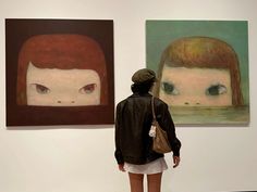 a woman standing in front of two paintings on the wall and looking at one another