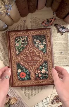 two hands are holding an ornate book on top of a table with books and butterflies