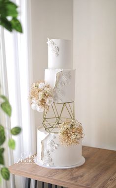 a three tiered white wedding cake with flowers on the top and sides, sitting on a table in front of a window