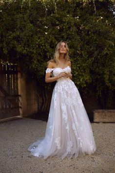 a woman in a white wedding dress standing next to a tree and bushes with her hands on her hips