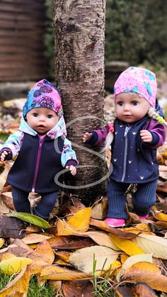 two small dolls are standing next to a tree in the leaves on the ground and one is wearing a hat