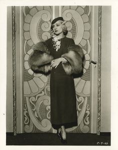 a black and white photo of a woman standing in front of a door wearing a fur stole