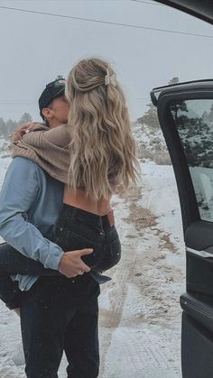 a man holding a woman in his arms while standing next to a car on a snowy road