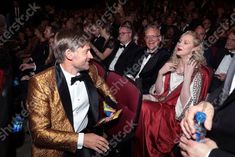 two men in tuxedos sitting next to each other at a formal event with people looking on