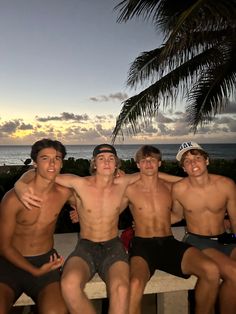 four young men sitting on a bench in front of the ocean and palm trees at sunset