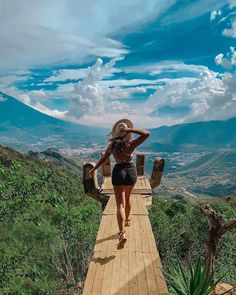 a woman walking across a wooden bridge over a lush green hillside under a blue sky