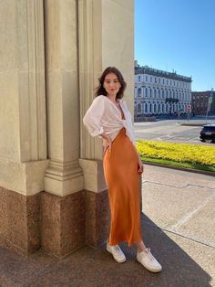 a woman standing next to a tall pillar in an orange skirt and white shirt with her hands on her hips