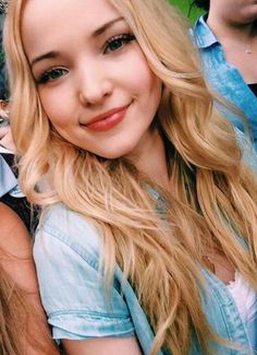 a woman with long blonde hair and blue shirt smiling at the camera while standing in front of other people