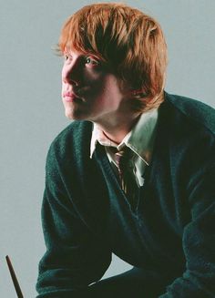 a young man with red hair wearing a sweater and tie, sitting in front of a white background