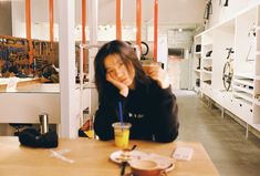 a woman sitting at a table with a drink in front of her and an empty shelf behind her