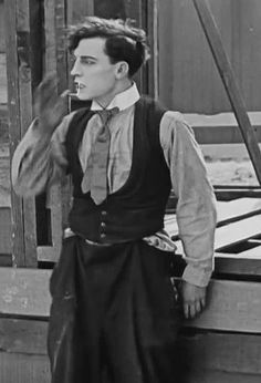 a man wearing a vest and tie standing in front of a wooden structure with his hand up