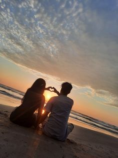 two people sitting on the beach at sunset making a heart shape with their hands as the sun sets