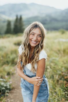 a woman in overalls is smiling while standing on a dirt path with mountains in the background