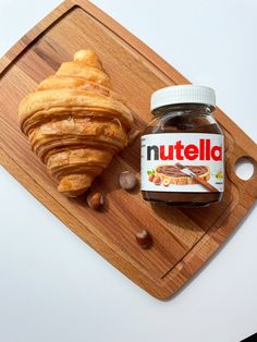 a wooden cutting board topped with a jar of nutella next to a croissant