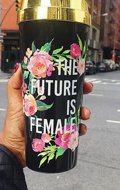 a person holding up a black and gold cup with flowers on it that says the future is female