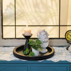 a candle and some plants on a tray with a clock in the background, sitting on a table