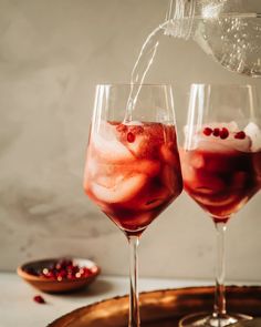 two wine glasses filled with red liquid being poured into one glass and topped with pomegranates