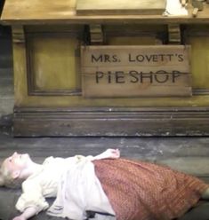 a woman laying on the ground in front of a wooden table with a sign that reads mrs lovett's pie shop