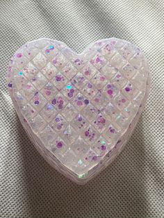 a white heart shaped box with pink and purple sequins on the inside sitting on a bed