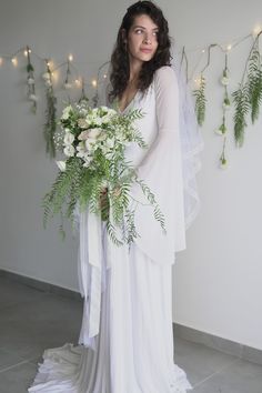 a woman in a white dress holding a bouquet of flowers and greenery with lights on the wall behind her