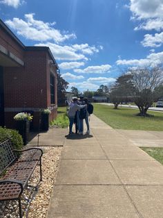 three people are standing on the sidewalk in front of a brick building with grass and flowers