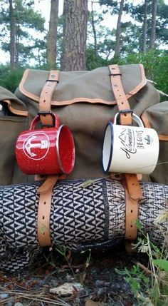 two coffee mugs sitting on top of a log in the woods next to a duffel bag