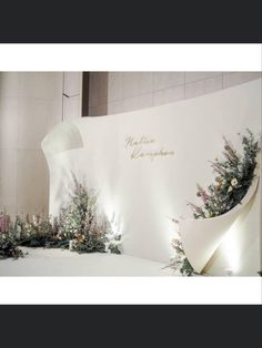 a white wall with flowers and greenery on it in front of a large sign