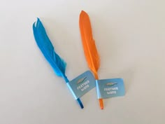 two blue and orange feathers are next to each other on a white surface with a name tag