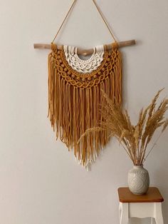 a white vase sitting on top of a wooden table next to a wall hanging with tassels