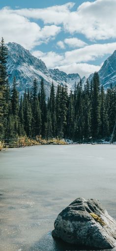 a large rock sitting in the middle of a lake surrounded by trees and snow covered mountains