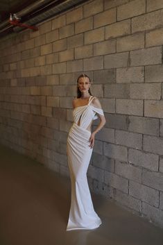 a woman standing in front of a brick wall wearing a white dress with one shoulder