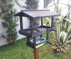 a bird feeder in the grass next to a house and planter with a cactus behind it