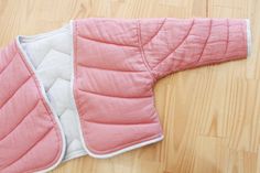 a pink and white quilted jacket laying on top of a wooden floor next to a pair of scissors