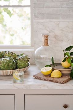 a counter topped with fruit and vegetables next to a vase filled with lemons on top of a wooden cutting board