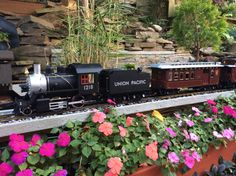 a toy train is on the tracks next to some flowers and plants in front of a stone wall