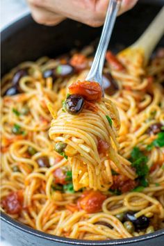 someone is holding a fork full of spaghetti with olives and tomatoes on it in a skillet