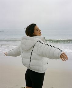 a woman is standing on the beach with her arms out and she is wearing a white puffy jacket