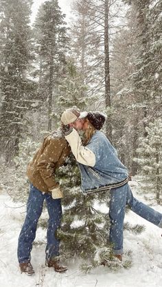 two people are kissing in the snow near a pine tree, with one person leaning on another