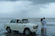 two people standing next to an old car in the water on a beach under a cloudy sky