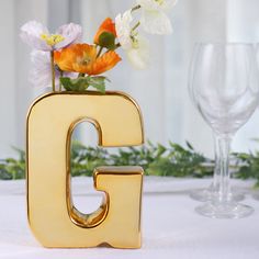 there is a vase with flowers in it and the letter g on the table next to two wine glasses