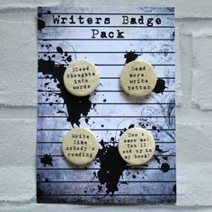 two white buttons with writing on them sitting on top of a wooden sign that says, writer's badge pack